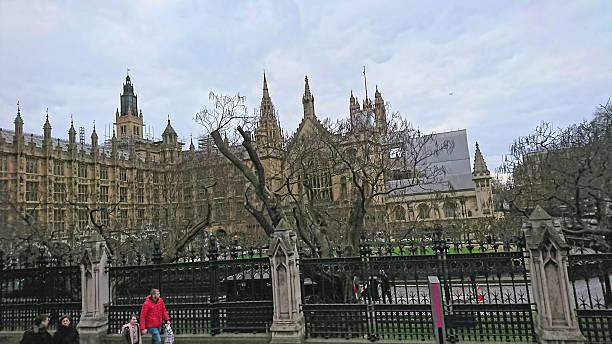 London, UK - February 18 2018: Pedestrians walk along the pavement next to the Palace of Westminster, home of the UK Parliament, with scaffolding visible on the Victoria Tower under an overcast