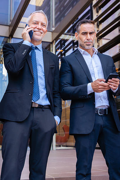 istock Two businessmen in suits using smartphones, talking outside modern office. 2247985222