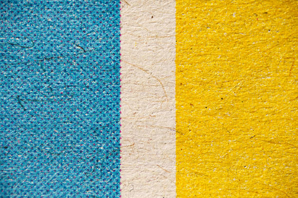 Extreme close-up of fibrous paper with vertical blue, beige, and yellow bands, showing halftone dots on the blue area and a bold flag-like stripe pattern.
