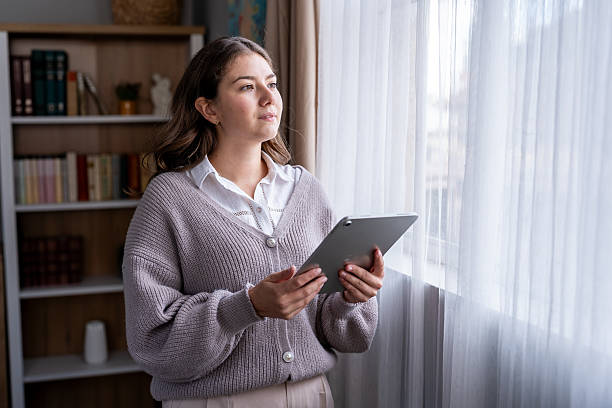 istock Young woman holding tablet looking out window at home 2247992733