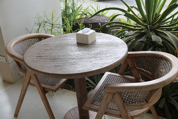 a round wooden table with two rattan chairs on the balcony, and an empty tissue box placed in front of it. the background is green plants and white walls, creating a fresh, natural style.