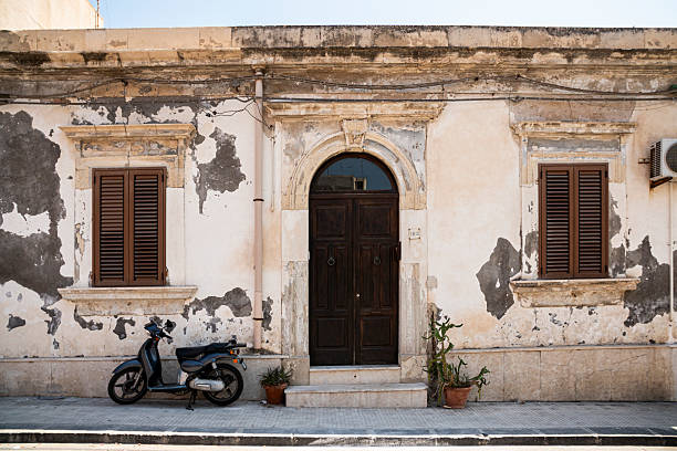 A dark grey scooter is parked in front of a traditional, weathered Mediterranean building in Sicily, likely in the historic city of Siracusa or nearby towns like Modica or Palermo. The rustic facade features an arched wooden door, two windows with brown wooden shutters, and classical stone surrounds, set on a simple street with potted plants, capturing the authentic charm of Southern Italian urban life.