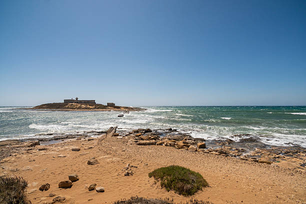 The image captures the rugged beauty of Isola delle Correnti (Island of the Currents), an islet off the coast of Portopalo di Capo Passero, Sicily. This unique location marks the geographical point where the Ionian Sea and the Mediterranean Sea converge, creating distinctive currents and a visually stunning natural spectacle. The foreground features a sandy beach and rocky shoreline, leading to the small island topped with an old, disused military structure and an active, solar-powered lighthouse. It is considered the southernmost point of the island of Sicily and a popular spot for nature lovers and photographers.