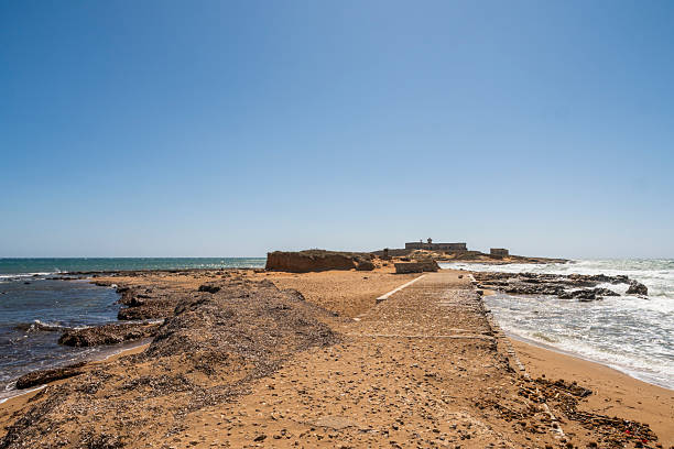 The image captures the rugged beauty of Isola delle Correnti (Island of the Currents), an islet off the coast of Portopalo di Capo Passero, Sicily. This unique location marks the geographical point where the Ionian Sea and the Mediterranean Sea converge, creating distinctive currents and a visually stunning natural spectacle. The foreground features a sandy beach and rocky shoreline, leading to the small island topped with an old, disused military structure and an active, solar-powered lighthouse. It is considered the southernmost point of the island of Sicily and a popular spot for nature lovers and photographers.