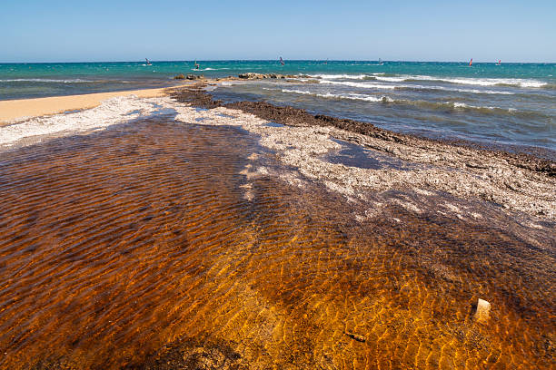 The image captures the rugged beauty of Isola delle Correnti (Island of the Currents), an islet off the coast of Portopalo di Capo Passero, Sicily. This unique location marks the geographical point where the Ionian Sea and the Mediterranean Sea converge, creating distinctive currents and a visually stunning natural spectacle. The foreground features a sandy beach and rocky shoreline, leading to the small island topped with an old, disused military structure and an active, solar-powered lighthouse. It is considered the southernmost point of the island of Sicily and a popular spot for nature lovers and photographers.