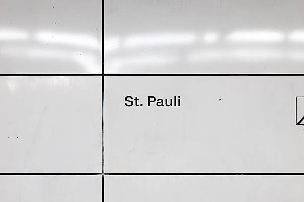 A close-up view of a St. Pauli bus stop sign in Hamburg, Germany, set against a background of leafy green trees and a clear blue sky.