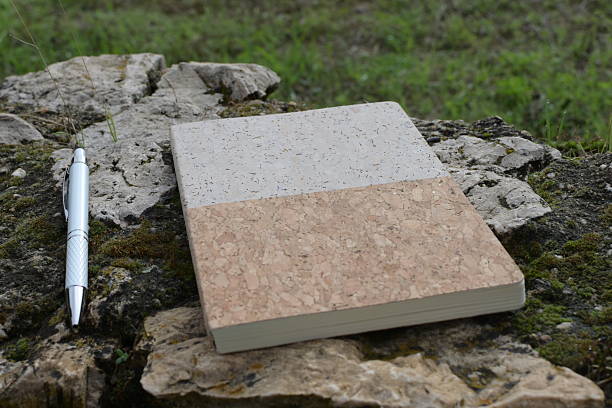 Textured notebook with a cork and stone-like cover sits beside a silver pen on rustic, moss-covered rocks, highlighting themes of nature, writing, and sustainable materials