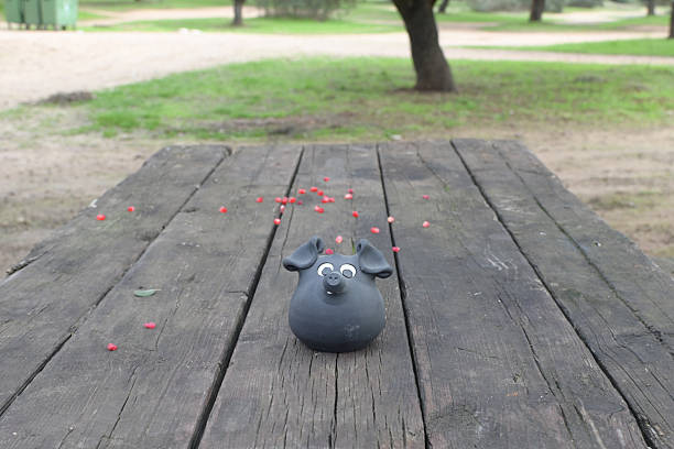 Black ceramic pig figurine with white eyes perched on a weathered wooden picnic table scattered with red berries in a quiet green park, soft natural light and shallow depth
