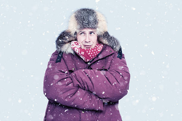 istock Man in red winter jacket and fur trapper hat shivers in falling snow, hugging himself for warmth against a cold, pale backdrop. Seasonal cold weather, freezing outdoor concept, harsh winter 2248026940