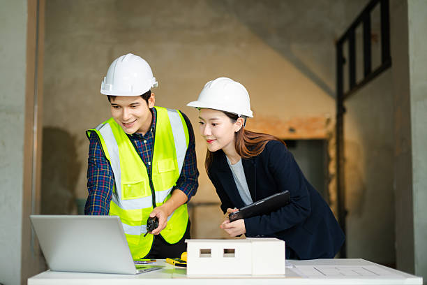 istock Construction engineer in a safety vest and an architect with a hard hat examine blueprints and a scale model on a table, collaborating 2248033517
