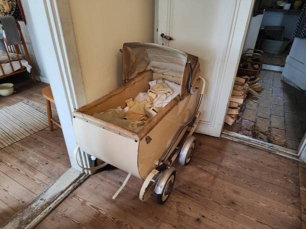 This photograph captures a vintage baby carriage placed indoors on light-colored wooden floorboards. The pram features a cream-colored body, metal frame, and four wheels with chrome hubcaps. Inside the deep carriage, various knitted blankets and baby clothes are visible beneath the raised hood. A partially open white doorway and stacked firewood are visible in the dimly lit adjacent room
