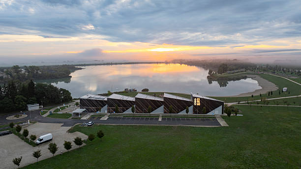 The Expano Pavilion stands beside a tranquil lake reflecting the dawn light, with a misty horizon in the background. The serene morning atmosphere creates a picturesque setting, capturing the beauty of nature and architecture in harmony.