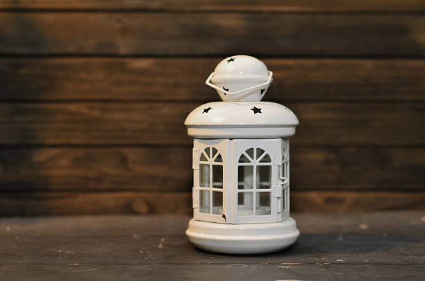 A vertical focus on a miniature white metal lantern candle holder featuring arched windows and star perforations on the roof, sitting on a dark surface and contrasted against a blurred horizontal dark