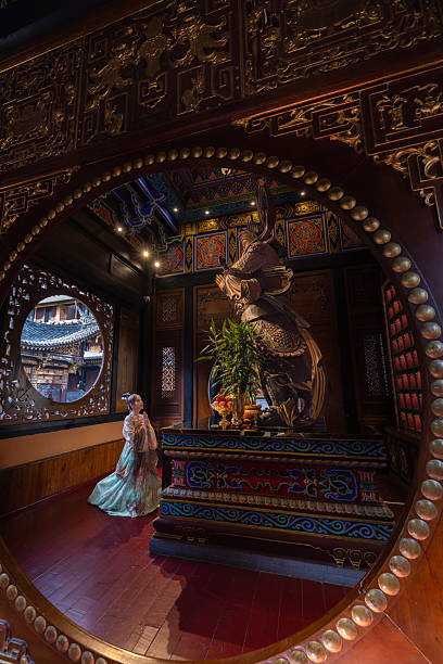 Luohan Temple, This captivating image captures a woman dressed in elegant traditional Chinese attire kneeling in prayer before a large, intricately carved deity statue inside an ornate temple. Sofe rays of light filter through the window. Chongqing , China.