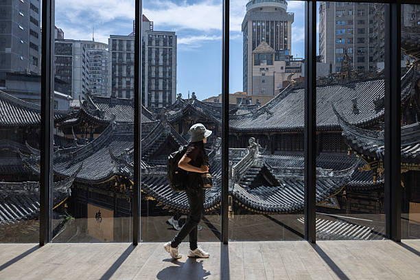A woman standing by a large window, gazing thoughtfully at the ornate rooftops of an ancient Luohan Temple complex below. Sunlight streams through the glass, creating a soft reflection and highlighting the contrast between the historical architecture outside and the modern city buildings rising in the background.