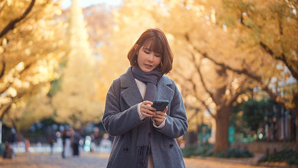 istock Young Woman Using Smartphone While Walking Through Autumn Ginkgo Tree Avenue in Tokyo. 2248054240