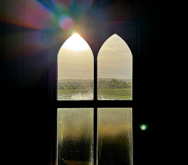 A view from a dark interior looking out through a four-pane window with a distinctive gothic arch design at the top. The sun is shining intensely through the upper left pane, causing a vibrant lens flare with rainbow light streaks to appear in the upper portion of the frame. A rural landscape can be seen through the windows.