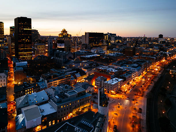 Aerial dawn view of downtown Montreal, showing illuminated skyscrapers, early morning city lights, modern office towers, dense urban streets and Mount Royal’s autumn foliage under soft sunrise light. Ideal for business, travel, real-estate and editorial use.