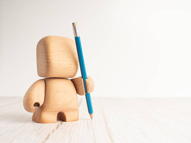 A stylized wooden toy figure is seen from the back, holding a blue pencil upright, on a pale wooden floor with a white background. Ideal for themes of creativity, education, or design.