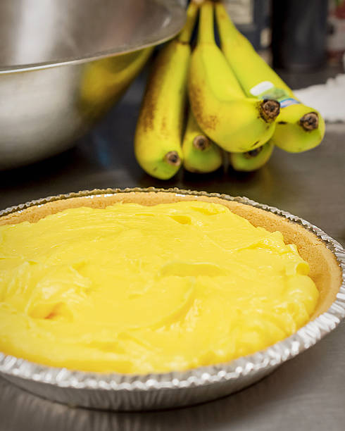 A finished layer of bright yellow banana cream pudding sits in a graham cracker crust on a stainless steel counter, with fresh bananas nearby in the cafe kitchen.
