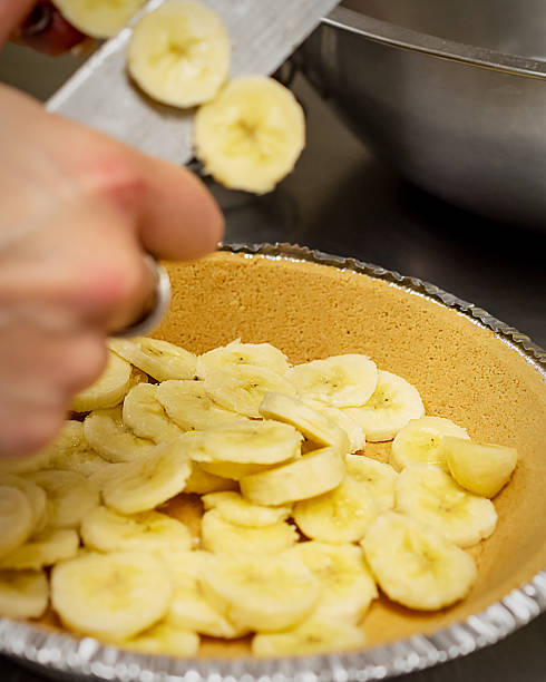 Close-up of a baker's hand slicing fresh bananas directly into a ready-made graham cracker pie crust, preparing the dessert base in a cafe kitchen.