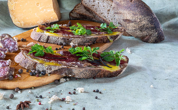 istock Artisan open-face sandwiches with cured meat and herbs on rustic bread, featuring a block of hard cheese and a loaf of bread in the background on a light linen tablecloth 2248089677