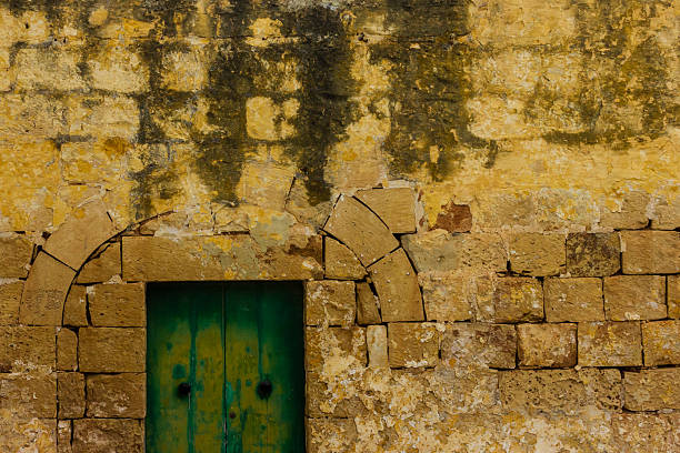 A weathered yellow stone wall frames a green wooden door with an arched stone surround, conveying rustic, historic charm and aged texture.
