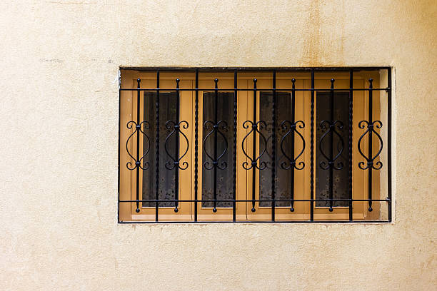A close-up of a rectangular window set in a sun-faded beige wall, protected by ornate black metal bars. The scene evokes urban security, rustic charm, and a calm, sunlit atmosphere.