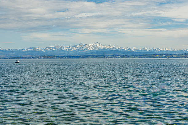 Lake Bodensee from the German side, with numerous sailboats floating on it. In the background is a panorama of snow-capped Alpine mountains. The photo was taken on a sunny day.