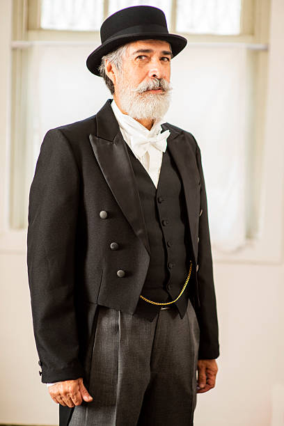 istock A mature man with a white beard, dressed in formal attire from the late 19th or early 20th century. He wears a top hat, black frock coat, waistcoat, white bow tie, and pocket chain, posing seriously in a well-lit setting. 2248096410