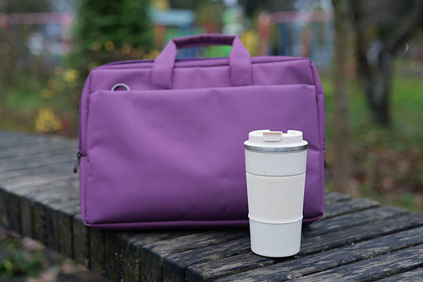 A purple bag sits next to a white coffee cup on a wooden bench in a park setting. The scene captures a peaceful afternoon ambiance with greenery
