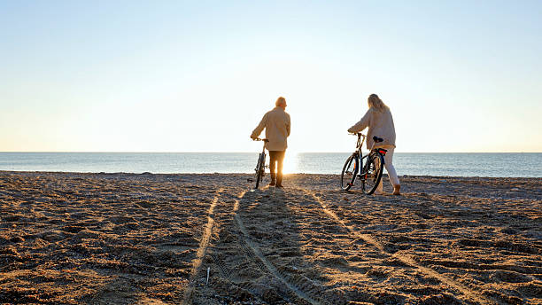 Mature couple push electric  bicycles onto beach at sunset, Liguria