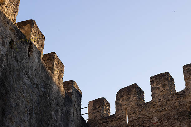 Lisbon, Portugal - May 31, 2019: The corner of São Jorge Castle’s walls rises sharply against a clear blue sky, with strong diagonal lines creating a sense of depth and perspective. The composition emphasizes the fortress’s imposing structure, giving the viewer an immersive feeling of being enclosed within the historic walls.