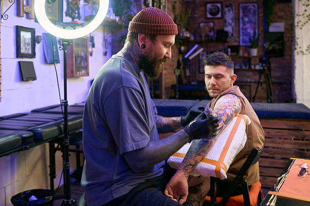 Caucasian young adult man tattoo artist working on forearm of Caucasian young adult man client sitting in tattoo studio, both focused on process, tattoo equipment visible in scene