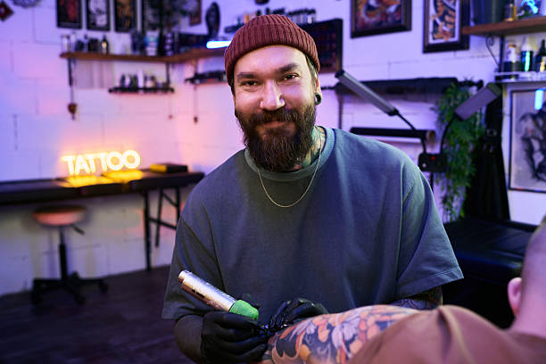 Portrait of young adult Caucasian man with beard smiling while tattooing client in modern tattoo studio, holding tattoo machine and wearing gloves, wall art and tattoo equipment visible in background