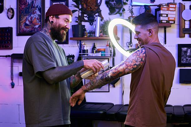 Caucasian young adult man with tattoos standing while another Caucasian young adult man with tattoos applying protective film to arm in tattoo studio