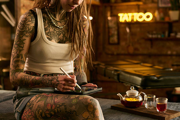 Young adult Caucasian woman with extensive tattoos sitting in tattoo studio using digital tablet with stylus, focusing on drawing design, teapot and cups on table nearby