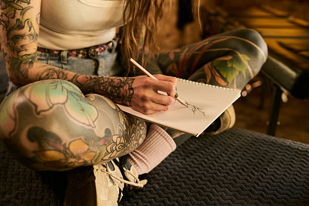 Caucasian young adult woman with long hair and extensive tattoos sitting cross legged on couch sketching in spiral notebook, holding pencil with left hand, focusing on drawing