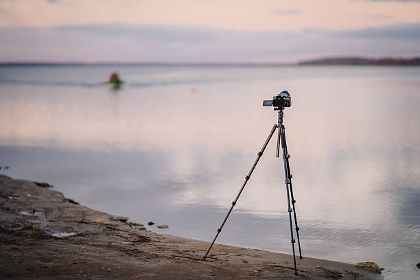Camera tripod positioned on rocky shore, capturing serene lake view at sunset, with gentle waves and distant silhouette of a person enjoying water activities