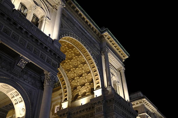 Night view of the Galleria Vittorio Emanuele II in Milan, showcasing its ornate archway and detailed neoclassical architecture beautifully lit against the dark sky. Warm artificial lighting highlights the sculpted decorations, columns and coffered ceiling, capturing the elegance of one of Milan’s most iconic landmarks.
