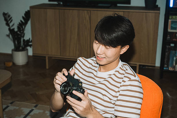 Medium shot of a focused young Asian man examining a vintage film camera while seated in an orange chair.