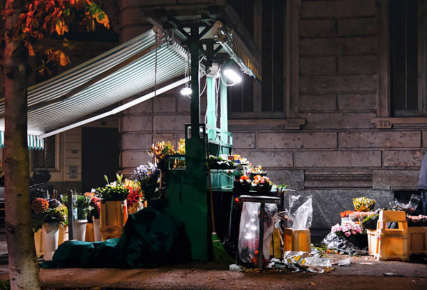 Colorful flower stall illuminated by bright artificial lights on a city sidewalk at night. Buckets of flowers, tools, and plastic wraps create a lively yet intimate urban market atmosphere.