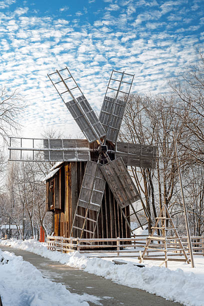 Rustic wooden windmill stands in a tranquil snow-covered field. A cleared path, wooden fence, and bare trees frame the scene beneath a bright blue sky with wispy clouds. The wintery setting conveys quiet, timeless rural charm and outdoor serenity.