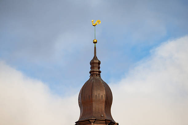 Tall spire in Riga rising from brown dome roof with distinct golden rooster element fixed to upper section and visible cloud formations in pale sky backdrop
