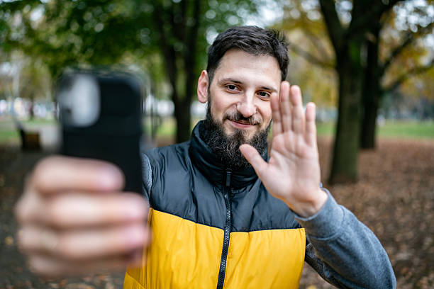 A bearded man in a yellow-and-black jacket takes a selfie in a park, smiling and waving at the camera as autumn leaves fall around him.