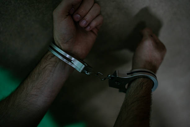 Dark, moody close-up of wrists in silver handcuffs linked together, capturing themes of arrest, confinement, and punishment. The dramatic shadows emphasize vulnerability and control in a tense moment.