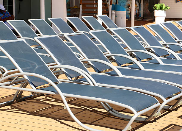 A sunlit, vivid blue ship deck features scattered metal chairs and tables while a lifebuoy hangs on the railing and the vast ocean stretches to the horizon, creating a peaceful nautical scene.