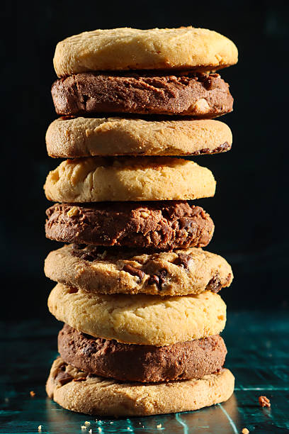Stock photo showing close-up view of a freshly made batch of homemade, chocolate chip biscuits stacked in tower on a mottled green surface, pictured against a black background. Home baking concept.