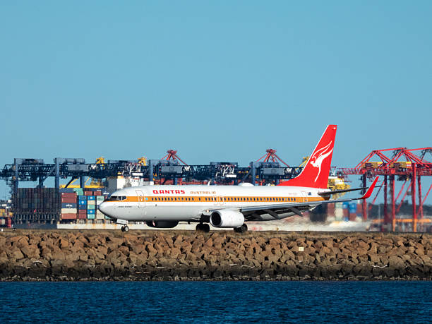 A Qantas Boeing B737-838 plane, registration VH-XZP in Retro Livery, taxiing to the international terminal of Sydney Kingsford Smith Airport after landing as flight QF164 from Wellington. In the background is the OOCL ITALY container ship docked at the Port Botany Container Terminal. This image was taken from near Kyeemagh Beach, Botany Bay on a sunny afternoon on 5 November 2025.