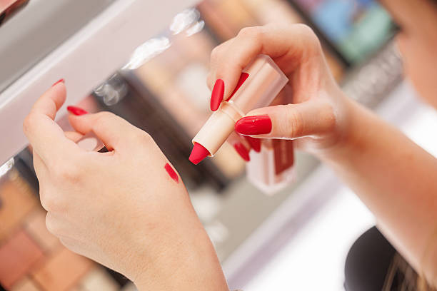 Manicured female hands trying on red lipstick on the back of hand in the make-up section by the illuminated shelf, shopping for beauty products